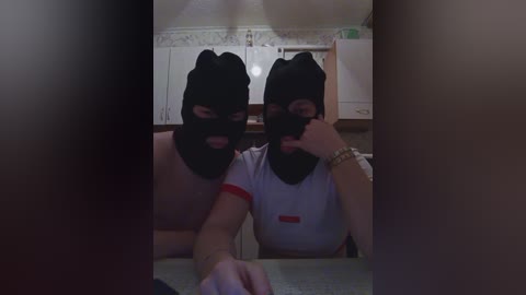 Video of two masked women in black hoods and white t-shirts, seated at a kitchen table, with a dimly lit, cluttered background featuring white cabinets and a microwave.