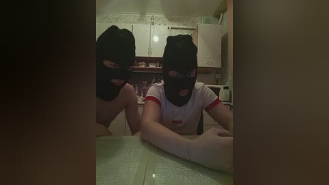 Video of two masked women in black balaclavas and red t-shirts, seated at a green table in a dimly lit kitchen with white cabinets and floral wallpaper.