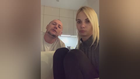 Media: Video of a man with a bald head and a woman with long blonde hair, both wearing casual attire, standing in a modern kitchen with beige cabinets and a white countertop.