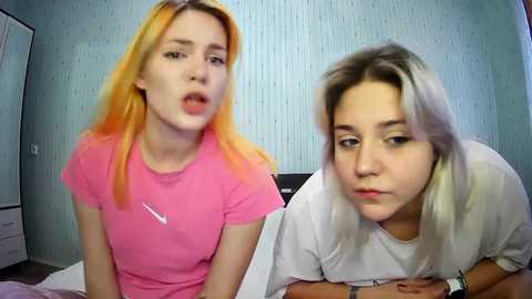 Media: Video of two young women with light skin and long hair, one in pink, the other in white, sitting on a bed, looking tired.