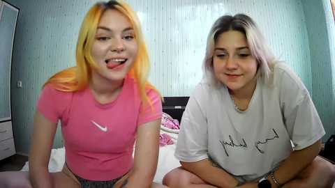 Media: Video of two young women with light skin and blonde hair, wearing casual pink and white t-shirts, sitting on a bed with white sheets, in a room with pale green wallpaper.