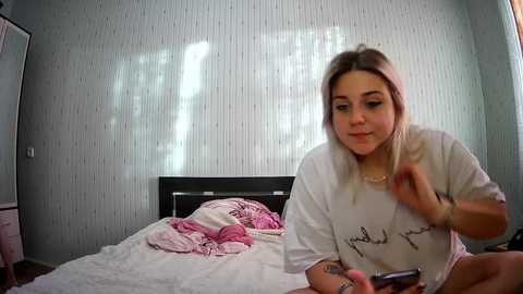 Media: Video of a blonde woman in a white T-shirt, sitting on a bed with pink sheets, holding a phone. The background features light grey, textured wallpaper and a window with blinds.