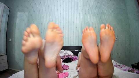 Media: Video of two pairs of feet with soles facing forward, lying on a bed with a pink and white striped blanket. The room has light blue, vertically striped wallpaper.