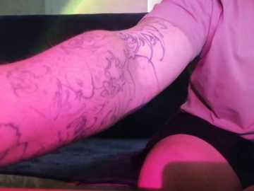 A close-up video of a person's arm with intricate black tattoo designs, wearing black shorts and a pink shirt. The background is blurred, with a soft pink glow, possibly indicating a night-time setting.
