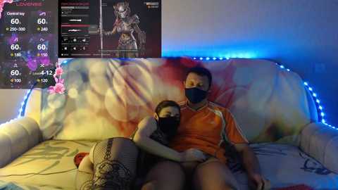 Video of an Asian man and woman on a couch, both wearing face masks, with a digital screen showing a character in a game.