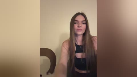 Media: Video of a young woman with long, straight brown hair, wearing a black sports bra, standing in front of beige walls, with a dark wooden chair to her left.