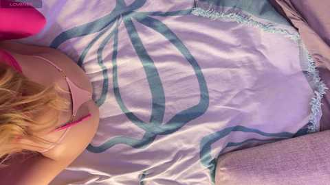 Media: Video of a blonde woman in a pink bra, lying on a bed with a white and teal patterned sheet.