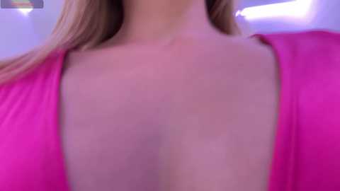 Media: A close-up video of a woman's bare chest with a prominent cleavage, wearing a vibrant pink, low-cut top. Her fair skin and long, blonde hair are visible. The background is blurred, featuring a mix of purple and blue hues.