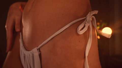 Media: Close-up video of a woman's lower back, wearing a white thong bikini, her skin glistening with oil, against a warm, dimly lit background.