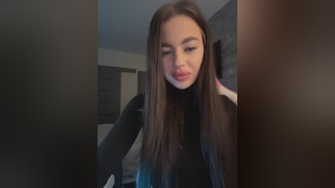 Video of a young woman with long brown hair, wearing a black top, slightly smiling, standing in a dimly lit bedroom with a grey wall and white dresser.