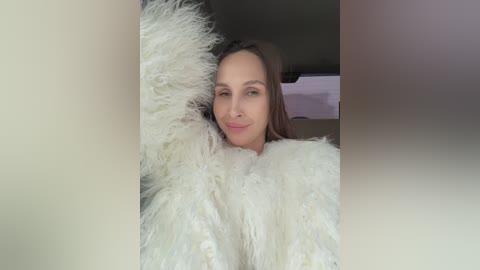 Video of a light-skinned woman with straight, medium-length brown hair, wearing a fluffy, white faux fur coat, smiling. The background is blurred, with a hint of a beige wall and a window.