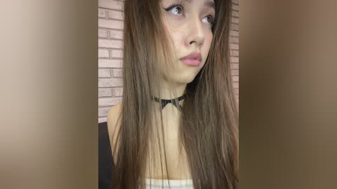 Video of a young woman with long, straight brown hair, wearing a black choker necklace and a black top, standing against a pink brick wall. She has a contemplative expression with her lips slightly parted.