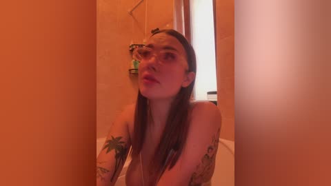 Video of a fair-skinned woman with long brown hair, wearing glasses, standing in a beige-tiled bathroom. She has a tattoo on her left arm, and the lighting casts an orange hue.
