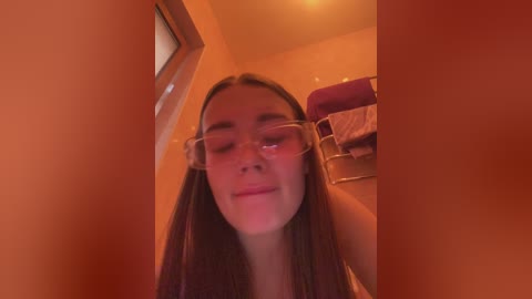 A video of a young woman with long, straight hair and light skin, wearing glasses and a white tank top, standing in a dimly lit bathroom with orange walls.