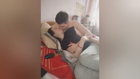 Video of two Asian men in a bedroom, one shirtless, lying on a bed, kissing, surrounded by red and beige blankets, wooden furniture, and a window.