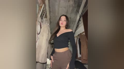 Media: A video of an Asian woman with long black hair, wearing a black off-shoulder top and high-waisted brown skirt, standing in a cluttered, dimly lit closet with hanging clothes and a concrete ceiling.