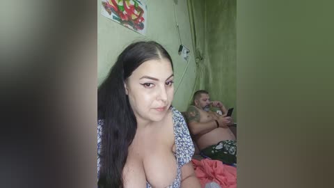 Video of a young woman with long black hair and large breasts, wearing a low-cut floral dress, in a dimly lit room with a green wall and a painting. A man in the background is shirtless, wearing patterned shorts.
