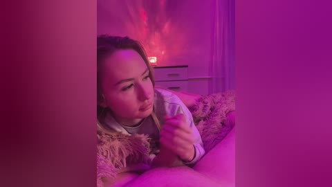 Video of a young woman with light skin and brown hair lying on a bed, holding a pink device, bathed in pink and purple ambient lighting.