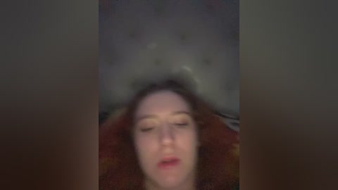 A blurred video of a person lying face up, eyes closed, with a light brown, furry blanket covering the upper body. The background is indistinct, with muted colors and a dimly lit, possibly indoor setting.