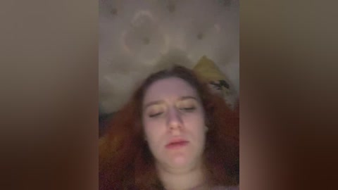 A blurry video of a woman with red hair, lying on her back, eyes closed, wearing a beige headpiece, against a dark background.