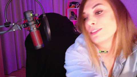 A video of a smiling, light-skinned, blonde woman wearing a blue shirt, sitting in front of a black chair and a purple-lit room with a red microphone stand.