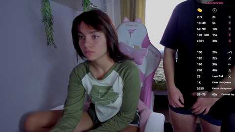 Media: Video of a young woman with long dark hair, green shirt, seated on a chair in a dimly lit room. A man stands behind her, partially visible, wearing a black shirt and shorts. A \"Love Live!\" poster hangs on the wall.