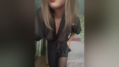 A video of a light-skinned woman with blonde hair, wearing a sheer black robe and black lace panties, leaning forward, her face partially obscured. Background features a green floral wallpaper and a bed with white sheets.