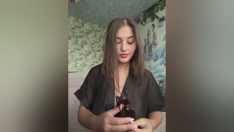 A video of a young woman with straight, shoulder-length brown hair, wearing a sheer black top, holding a pair of dark sunglasses in a bedroom with floral wallpaper and a mural of a castle.