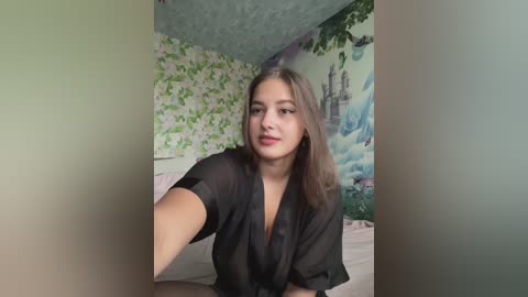 Video of a young woman with light skin, straight brown hair, and brown eyes, wearing a black sheer top, sitting on a bed in a room with green floral wallpaper and a mural of a castle on the wall.