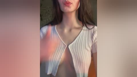 Video of a woman with fair skin and long dark hair, wearing a white ribbed cardigan with a plunging neckline, partially unbuttoned, showcasing cleavage. Background features blurred abstract art.