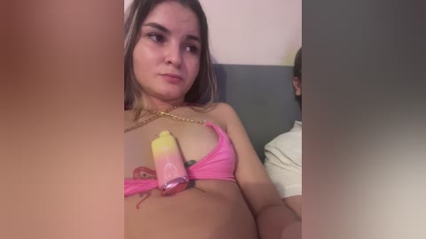 Video of a young woman with light skin, long blonde hair, wearing a pink bikini, holding a colorful popsicle, and sitting on a bed in a dimly lit room.