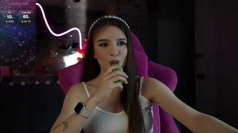 Media: Video of a young woman with long brown hair, wearing a white camisole and a pink headset, eating a green apple in a dimly lit room with a neon sign in the background.