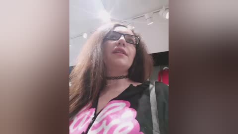 Video of a young woman with long brown hair, wearing black-framed glasses, a black choker, and a black hoodie with pink flamingos. The background is a dimly lit, cluttered room with clothes hanging.