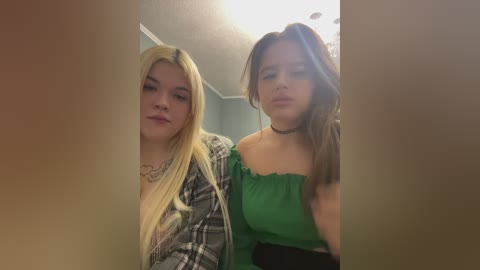 Video of two young women with long blonde and brunette hair, wearing plaid shirts and green tops, standing closely in a dimly lit room.