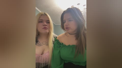 Video of two young women with long blonde and brown hair, respectively, in a green off-the-shoulder dress and a pink fringed top. They stand indoors near a chandelier.