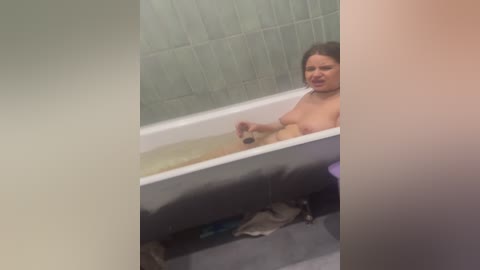 A video shows a nude woman with light skin, medium-sized breasts, and dark hair, sitting in a bathtub filled with water, in a bathroom with tiled walls and a purple bath mat.