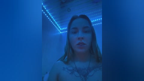 Media: Video of a young woman with long, wet, blonde hair, wearing a white lace bra, standing in a dimly lit room with blue LED strip lighting on the ceiling.