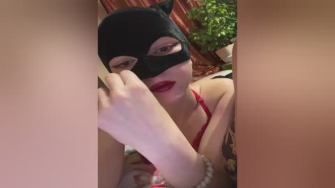Video of a woman in a black Catwoman mask, red lipstick, and a red bra, holding her bare foot forward. Background shows green plants and curtains.