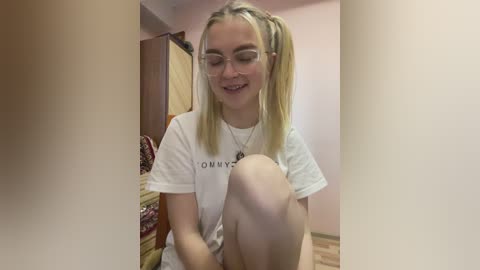 Video of a young, smiling woman with straight blonde hair in pigtails, wearing glasses and a white t-shirt. She is sitting on a bed with wooden floors and a patterned blanket in a room with light pink walls.