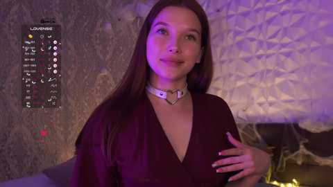 A video of a smiling, light-skinned woman with long brown hair, wearing a black dress and white choker, in a dimly lit room with purple lighting and a textured wall.