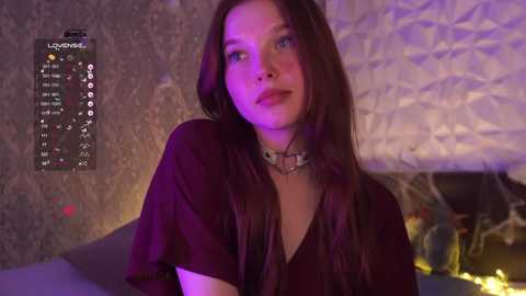 Video of a young woman with long dark hair, wearing a dark blouse and a choker, in a dimly lit room with purple and yellow lighting.