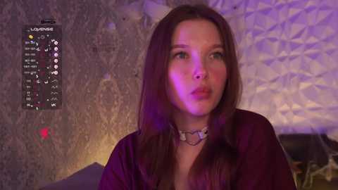 Video of a fair-skinned woman with long brown hair, wearing a maroon top, against a textured, purple-lit wall. She gazes intently with a neutral expression.