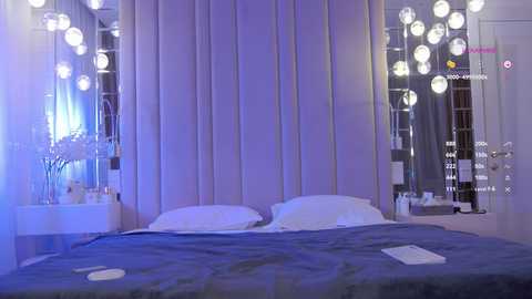 Video of a modern bedroom with a white tufted headboard, white pillows, and a blue blanket on a bed. Surrounding the bed are spherical, white hanging lights.