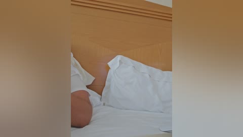A video of a messy bed with white pillows and a light brown headboard, partially obscured by a large, beige object in the foreground.