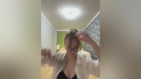 Media: Video of a woman with glasses, brown hair tied in a bun, wearing a white shirt and black bra, adjusting her hair in a hallway with patterned wallpaper and a green curtain.