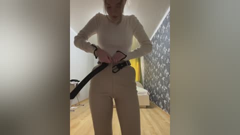 Media: Video of a slender, fair-skinned woman with long blonde hair, wearing a white long-sleeve shirt and high-waisted, tight beige pants, adjusting a black belt in a dimly-lit room with wooden floors and patterned wallpaper.