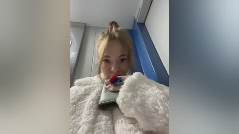 Media: Video of a young Asian woman with light skin, wearing a fluffy white coat and holding a green purse, looking directly at the camera. She has a high ponytail and is indoors with blue and white walls.