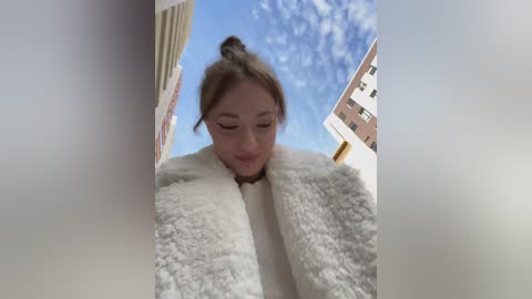 Media: Video of a young woman with light skin and brown hair in a messy bun, wearing a fluffy white coat, looking down at a camera from a high angle. Background features blue sky and modern apartment buildings.