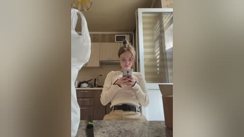 Media: A video of a young Caucasian woman with blonde hair taking a mirror selfie in a small, tidy kitchen with beige walls and wooden cabinets. She's wearing a beige sweater.