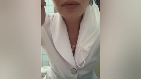 Media: Video of a woman in a white lab coat, wearing a gold necklace, in a bathroom with tiled walls, partially visible.